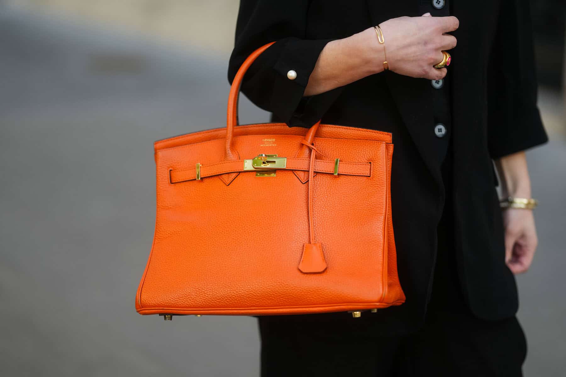: An orange Hermes Birkin bag in grained leather with structured handles, gold-toned hardware, a signature front lock, and tonal stitching is carried. A black blazer with pearl button cuffs is worn. A gold cuff bracelet and a sculptural gold ring with red enamel detailing.2025 in Paris, France. Picture Edward Berthelot/Getty Images