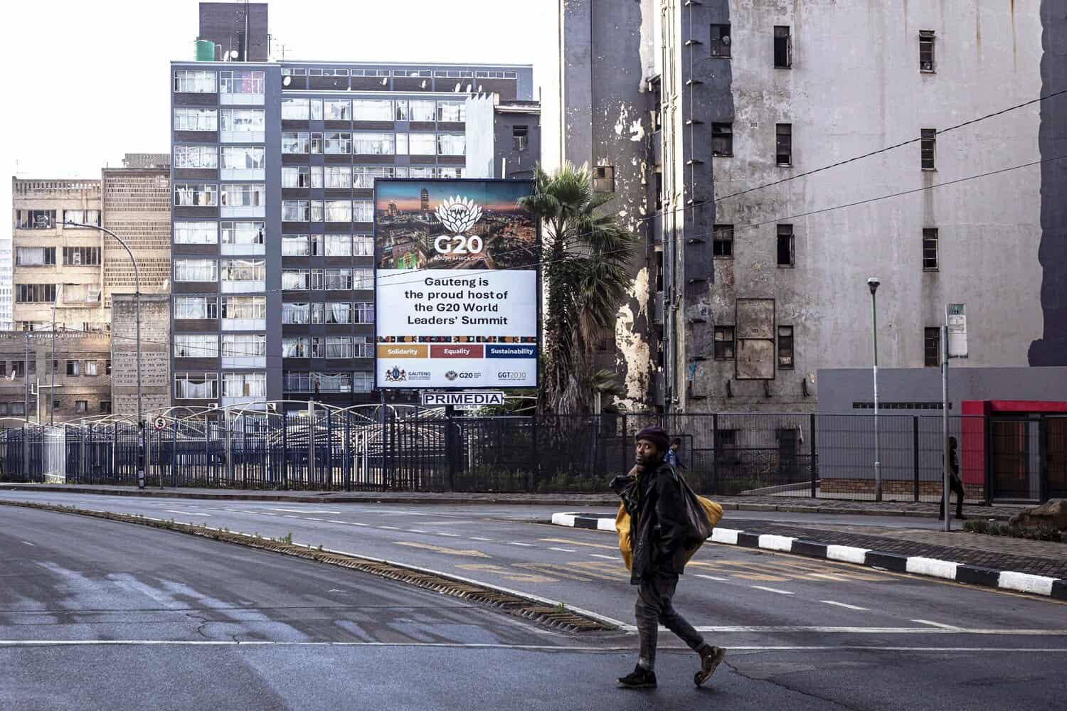 A homeless man walks past a billboard promoting the upcoming G20 Leaders's Summit stands among housing projects on November 8, 2025 in Braamfontein in central Johannesburg, South Africa.