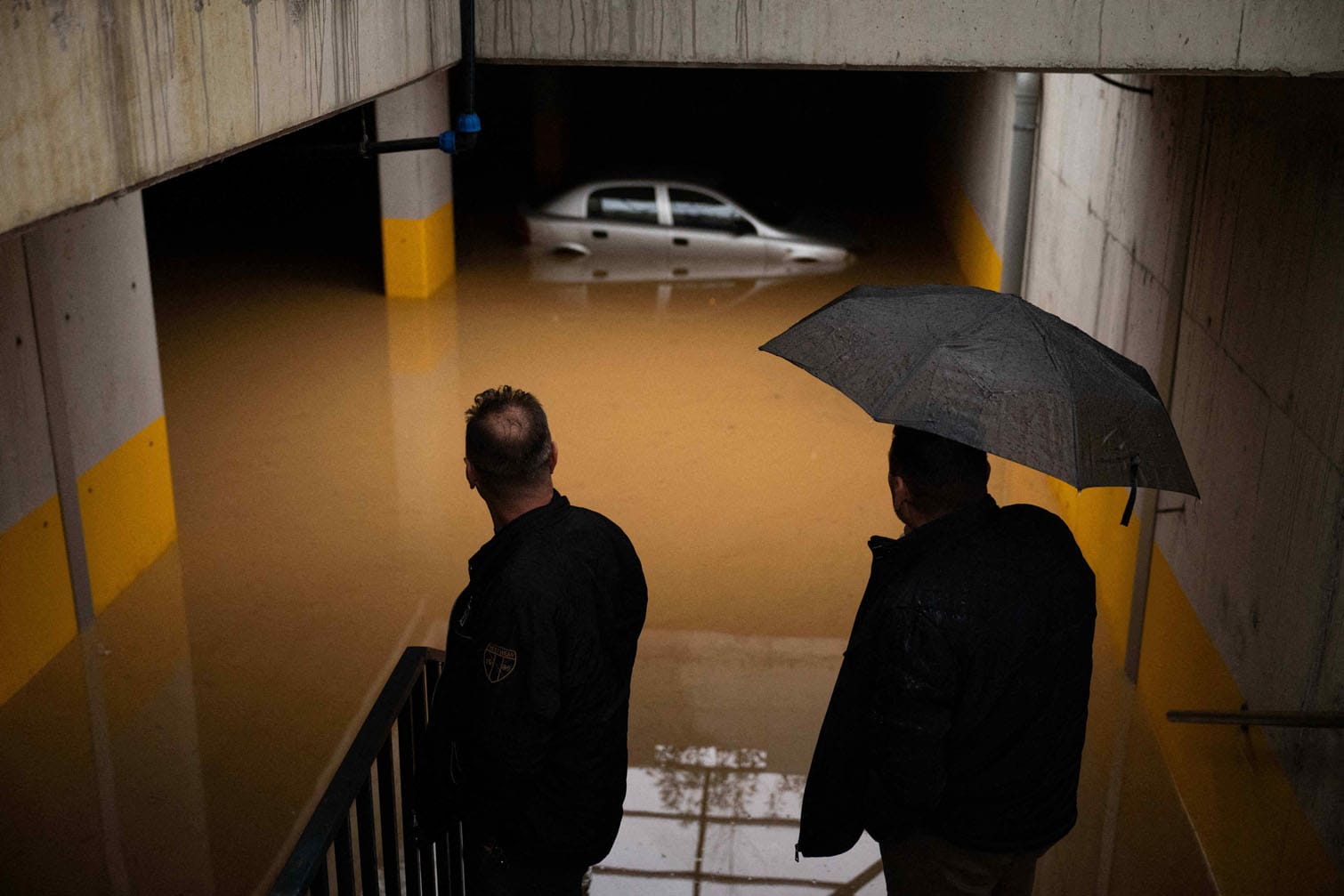 Residents look at a car submerged in a flooded parking lot