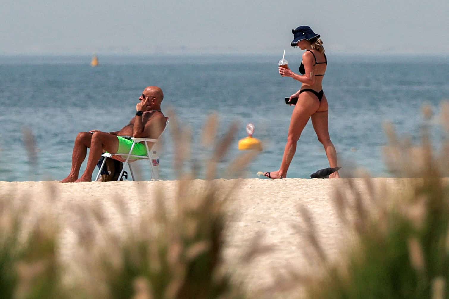 A woman walks with a drink near a man sitting along the beach in Dubai
