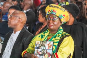Mama Joy at a public event, dressed in colorful traditional attire, holding photos, with a lively cr.