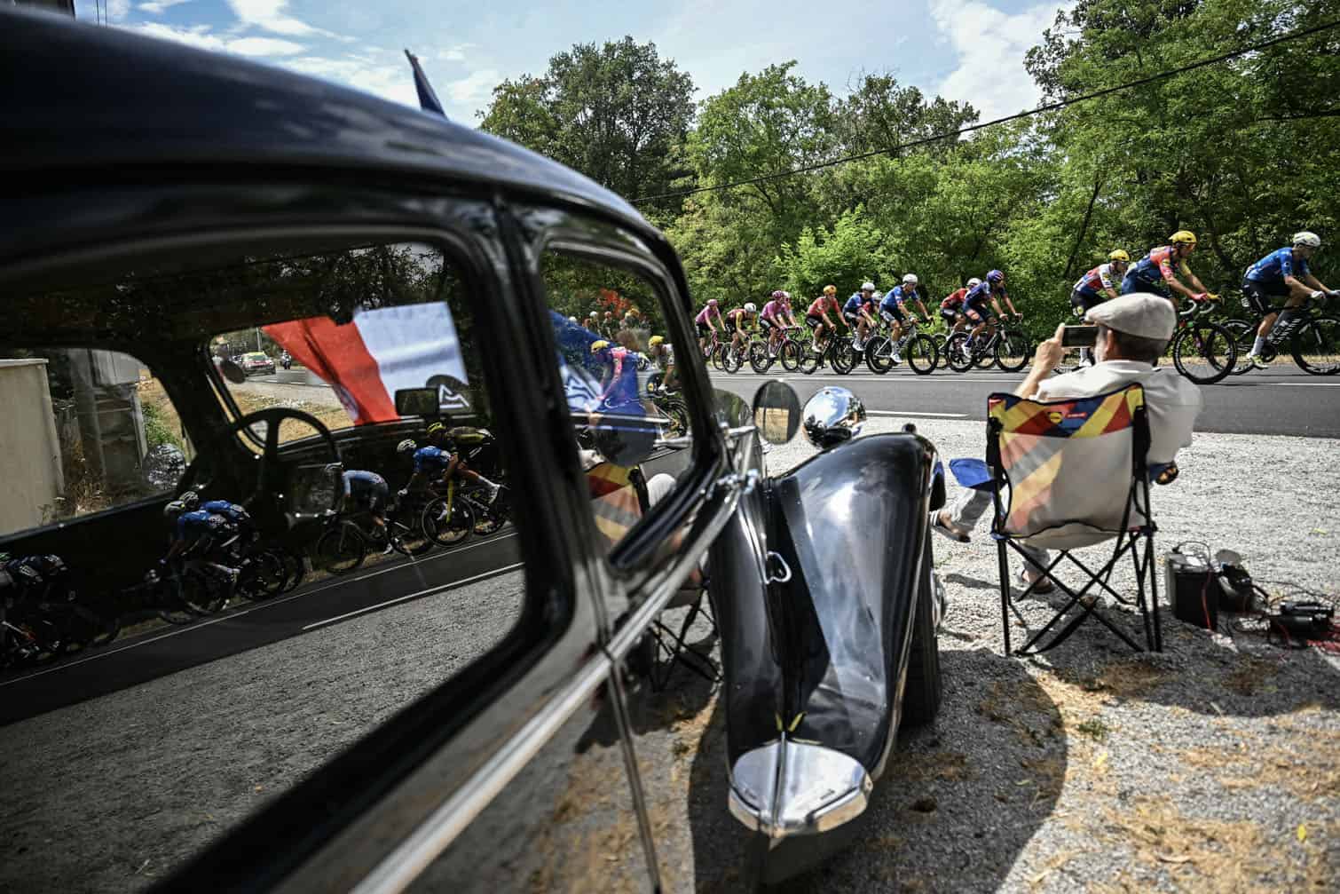 The pack of riders (peloton) cycles past a classic car