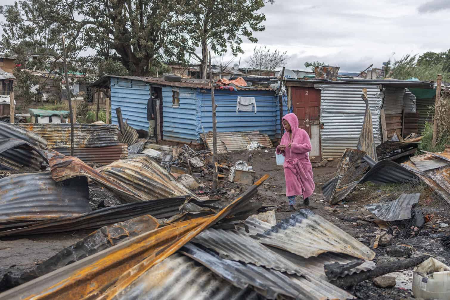A woman walks past structures damaged by a fire that destroyed more than 500 dwellings at the Jika Joe informal settlement in Pietermaritzburg, KwaZulu-Natal, August 11, 2026. The fire broke out on August 9, displacing more than 600 people during a severe cold front that brought freezing temperatures and disruptive snowfall to parts of the province. Survivors were left stranded without shelter, food or blankets and dependent on non-governmental organisations for assistance. (Photo by RAJESH JANTILAL / AFP)