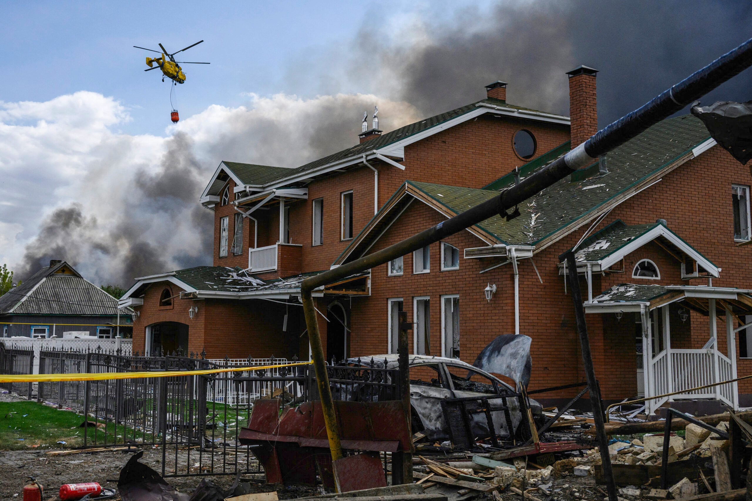 This photograph shows a damaged house as a firefighting helicopter carries a load of water to extinguish a fire at a warehouse following an air attack near Boryspil, Kyiv region on August 27, 2026, amid the Russian invasion of Ukraine. (Photo by Vladyslav Musiienko / AFP)