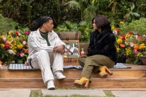 "Two people sit on a picnic blanket outdoors, facing each other and talking, surrounded by colorful flowers and greenery, with a picnic basket and food between them in a lush garden setting."