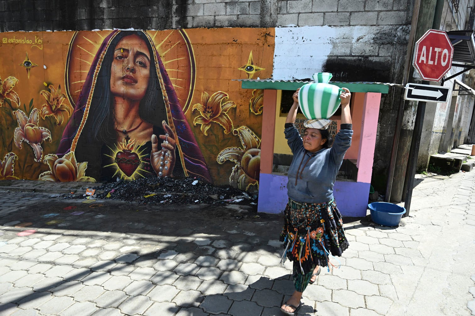 An Indigenous woman walks carrying plastic water containers as she passes a mural of the Virgin of the Sacred Heart of Jesus on one of the streets of Santa Maria de Jesus, Guatemala on September 30, 2026. The murals are part of “"A New Air for Santa Maria de Jesus" an initiative that seeks to restore deteriorated spaces and strengthen the identity of this community through art. (Photo by Johan ORDONEZ / AFP)