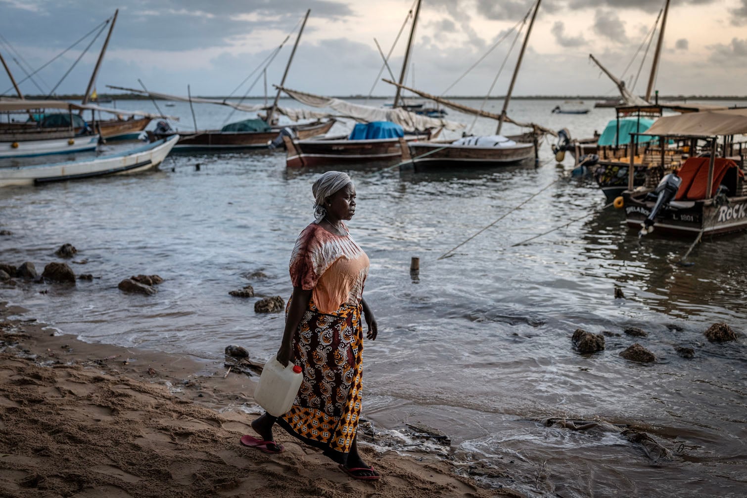 A woman walks along the shore at dawn as boats lie moored at a jetty on Lamu Island on October 1, 2026.The picturesque Kenyan island of Lamu faces a tough trade-off after becoming the construction site for Africa's latest mega-refinery.Lamu, on the Indian Ocean coast, is known for its 12th-century Old Town, traditional dhow sailing boats and paradisical beaches that have made it a favourite haunt of stars like Mick Jagger, Kate Moss and the Obamas. But within less than half a year it has become the chosen location for Africa's second-biggest oil refinery, being built by Nigerian tycoon Aliko Dangote. (Photo by Luis TATO / AFP)